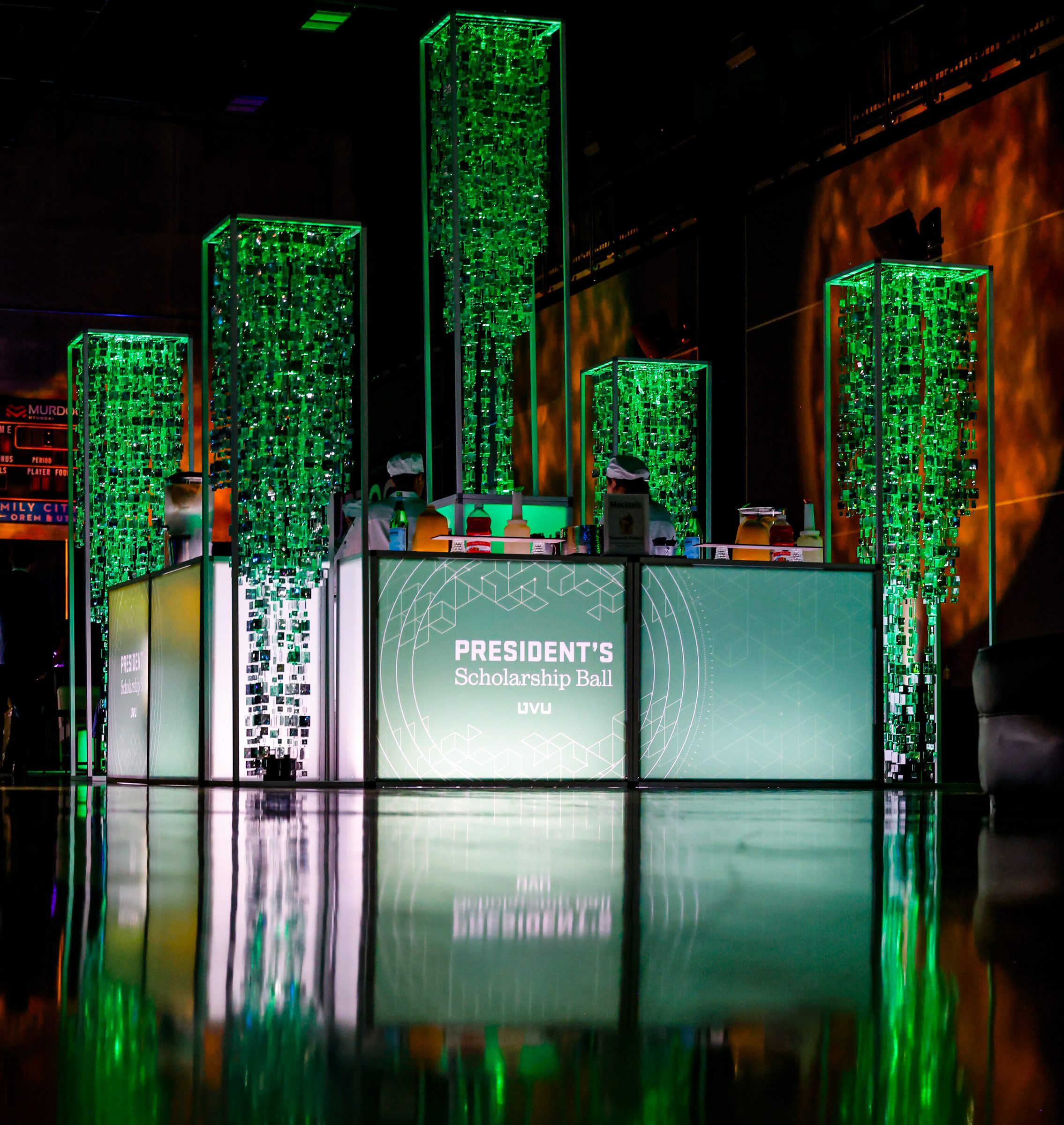 Backlit bar front with the President's Scholarship Ball lockup and lens rings, under hanging green crystal towers