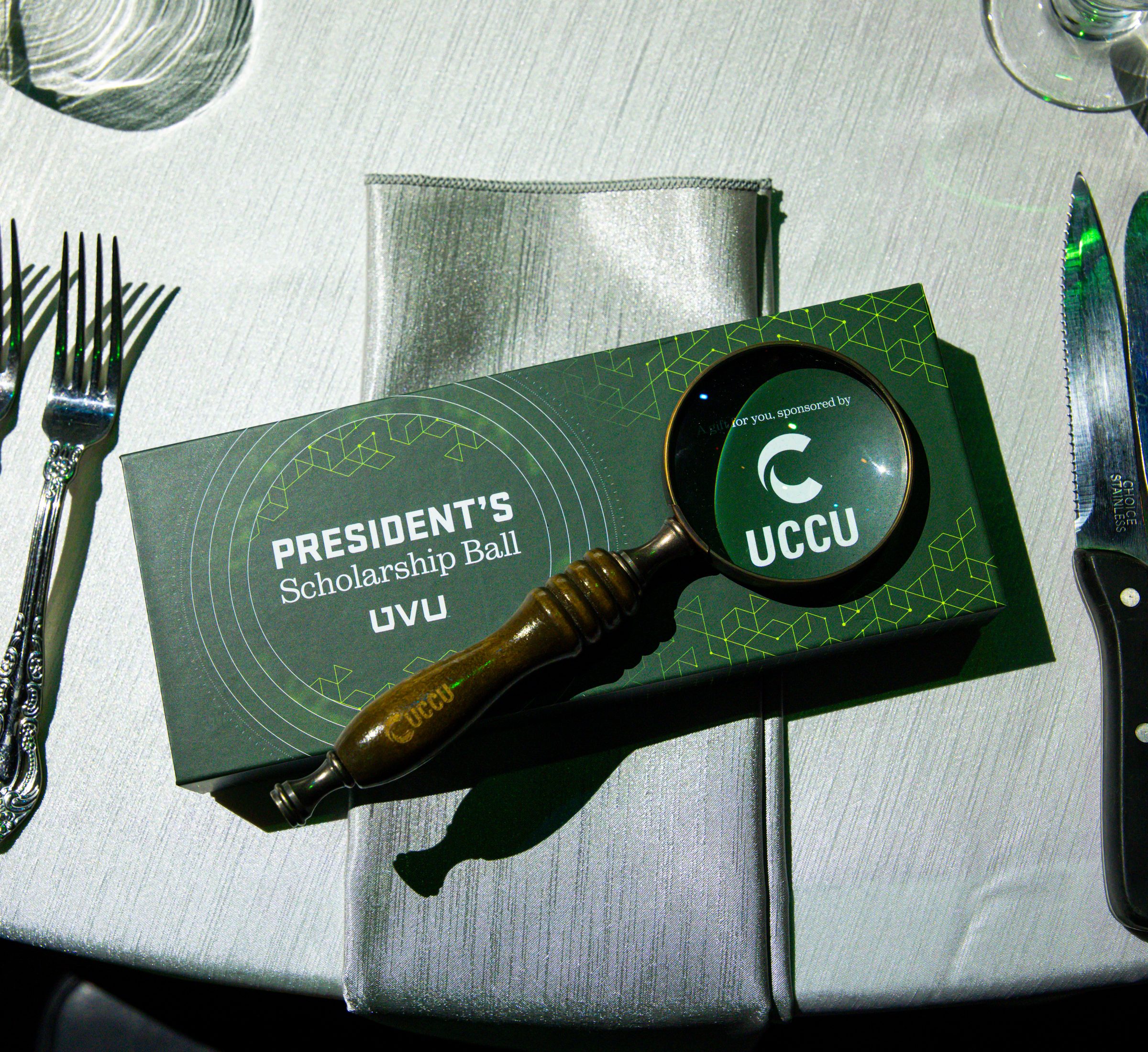 Green donor gift box with the PSB lockup and a brass magnifying glass framing the UCCU sponsor mark