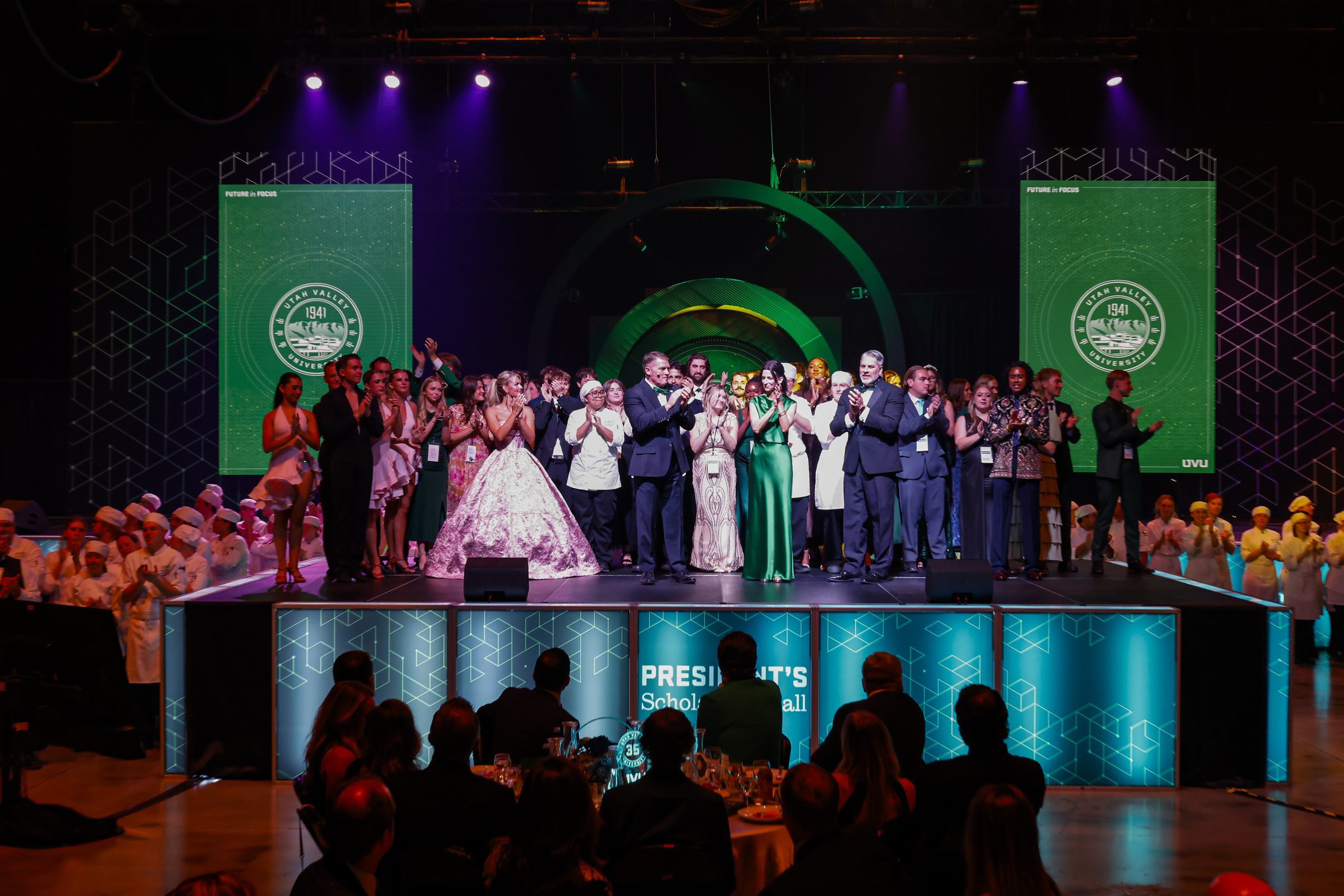 The full PSB 2026 cast, dancers, chefs, and students, applauding on stage between the Future in Focus screens and the glowing focal ring, above the President's Scholarship Ball stage front